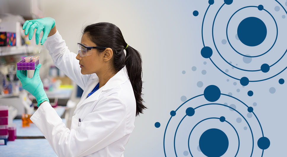 Image: Woman with dark hair wearing white lab coat and safety goggles examining a clear liquid in a small vial. Vial is held over a group of similiar vials in a purple holder.